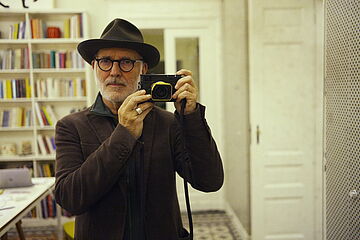 Ludovico mit Hut und einem Fotoapparat in den Händen steht in einem Zimmer mit einer Bücherwand. 